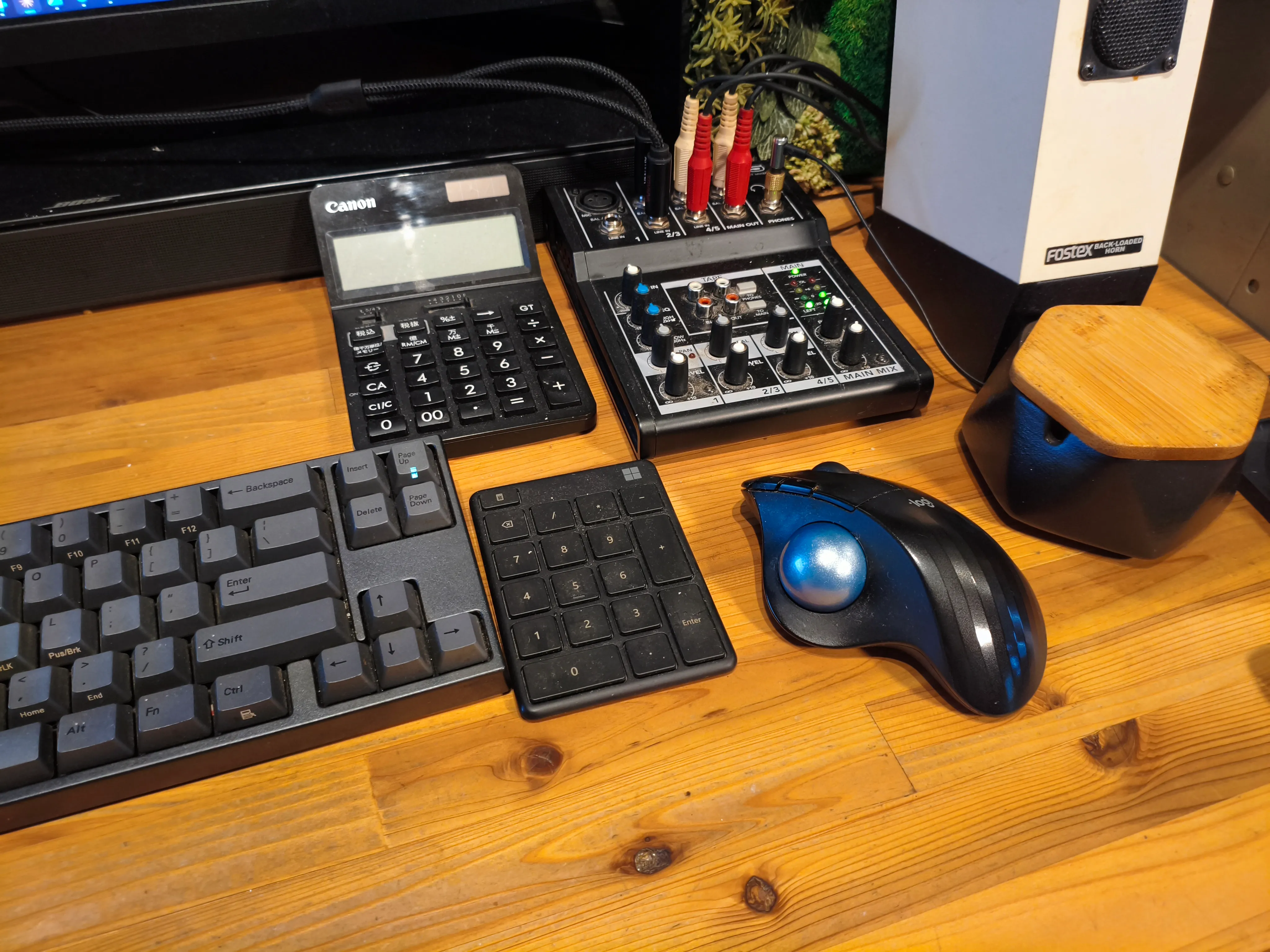 木製デスクに並んだ黒いキーボード、テンキー、電卓と、右手前に置かれた黒いトラックボール。マウスを滑らせる空きスペースはない