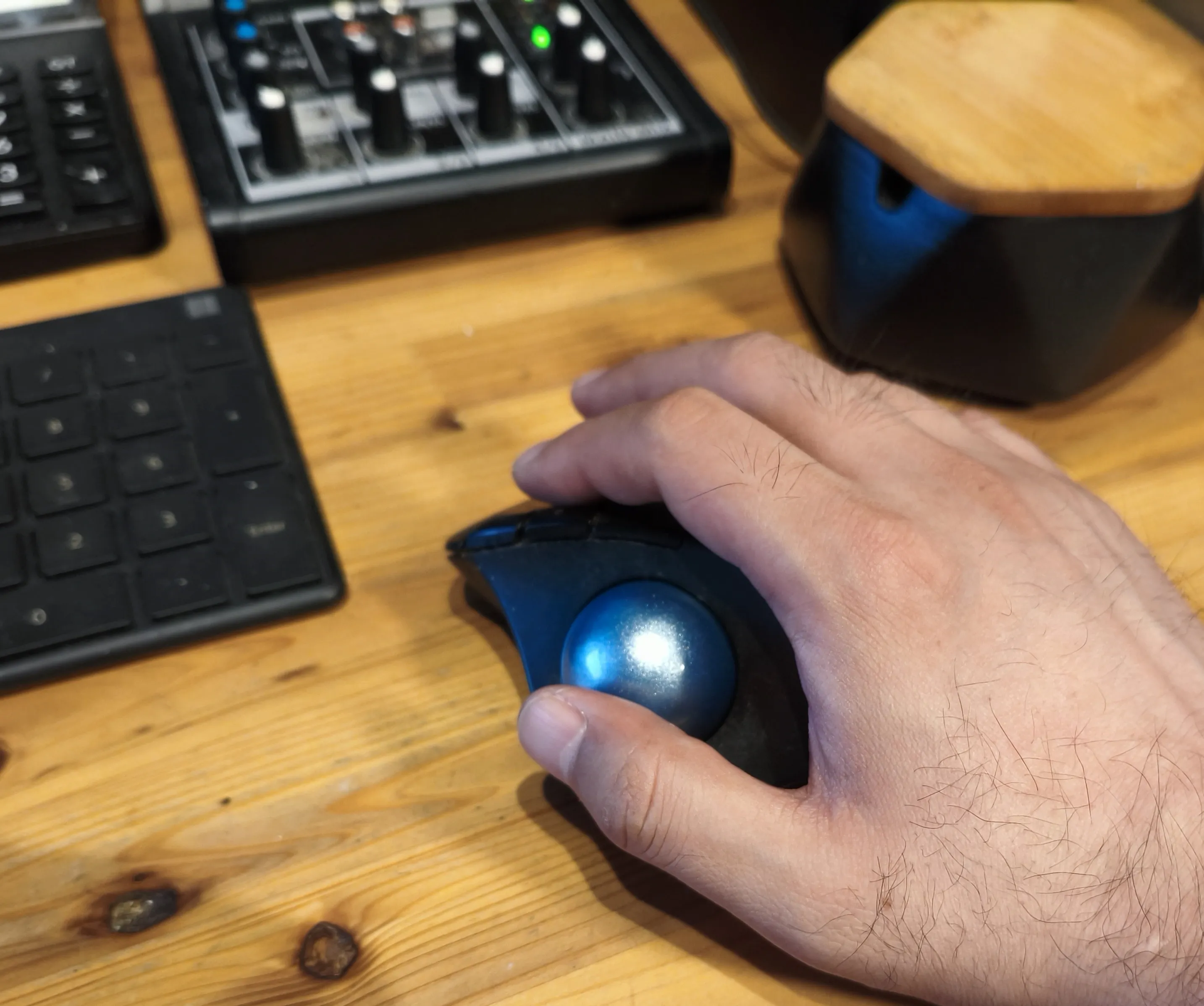 木製デスクの上で、黒いトラックボール型マウスの青いボールを親指で操作している手元。左にはテンキー、奥に黒い機材が見える